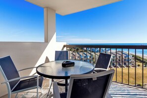 A balcony with a glass table and chairs, overlooking a beach and a row of colorful buildings. - 6th-Floor Oceanview Condo w/ Beautiful Ocean & Sunrise Views (Emerald Isle)