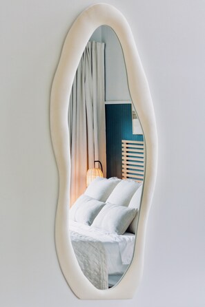 Ein Spiegel mit unregelmäßig ovalem Rahmen, der ein Bett mit Kissen und einer Nachttischlampe widerspiegelt.
