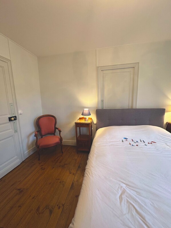 A hotel room with a wooden floor, a single bed, a chair, a nightstand with a lamp, and a door. - Maison Dellac – Les clés du temps (Saint-Saturnin)