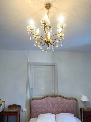Ein Zimmer mit einer Kandelaber, einem Bett mit einem gemusterten Kopfteil, einem Nachttisch und einer Tür.