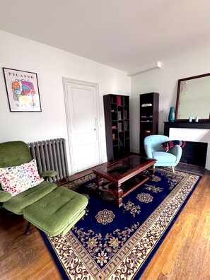 Un salon avec un canapé vert, un fauteuil bleu, une table basse en bois, un tapis à motifs, une bibliothèque et un tableau encadré accroché au mur.