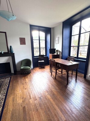 A room with a wooden dining table, a green armchair, a small stool, a television on a stand, and a large window with a view of a stone building. - Maison Dellac – Les clés du temps (Saint-Saturnin)
