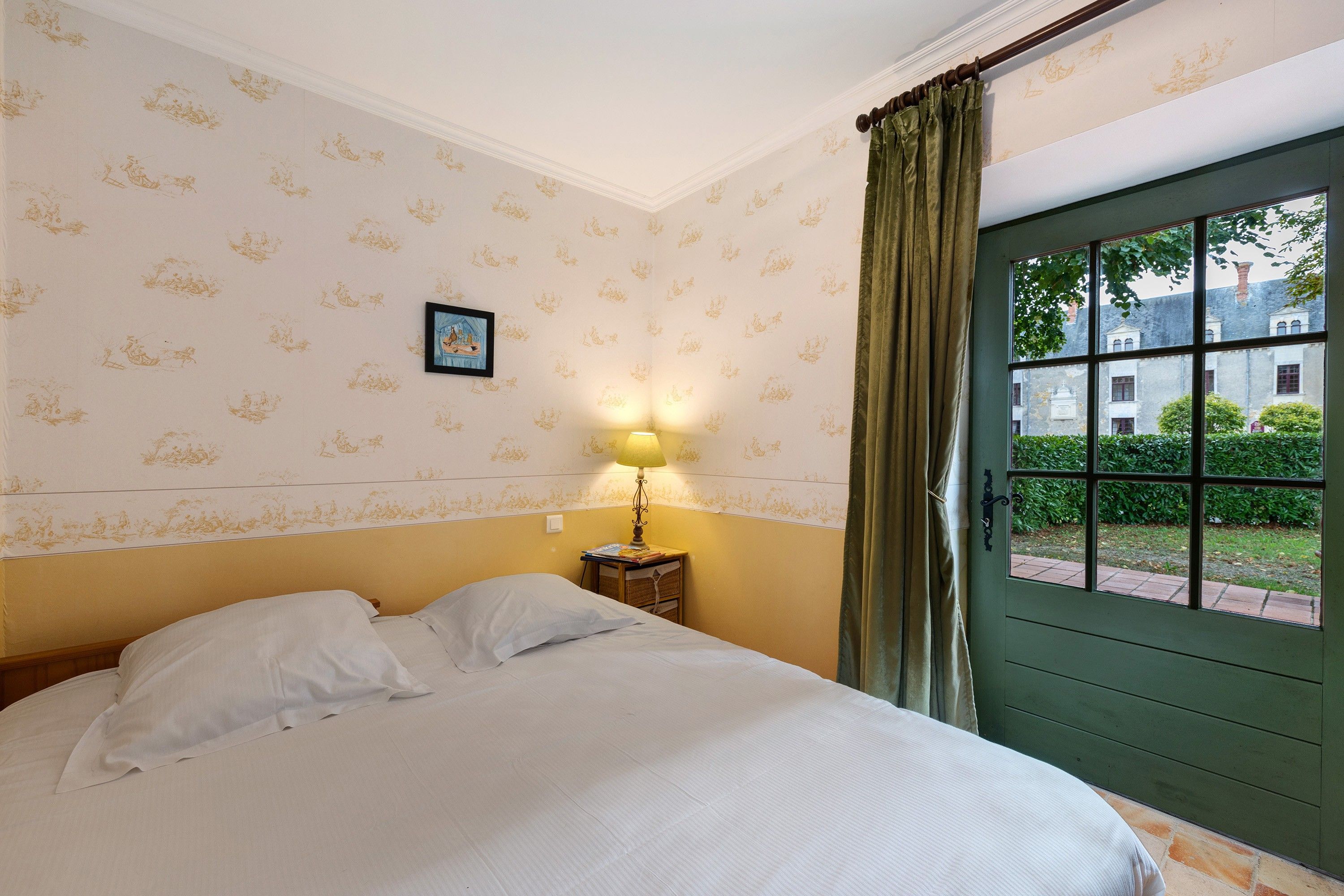 A bedroom with a bed, a bedside lamp, a framed picture, and a window with curtains.