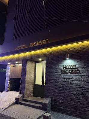 Entrada de hotel com placa indicando 'HOTEL PICASSO' e fachada de tijolos.