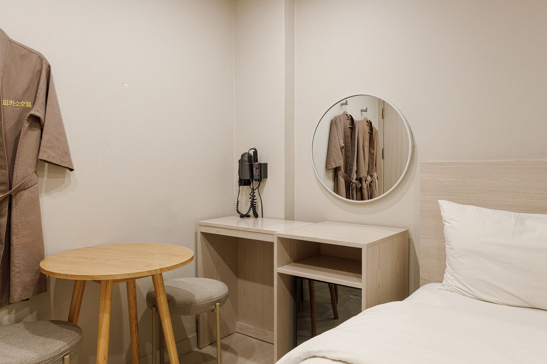 Una habitación de hotel con una cama, un espejo redondo, una mesita pequeña, un taburete y una bata colgada en la pared.