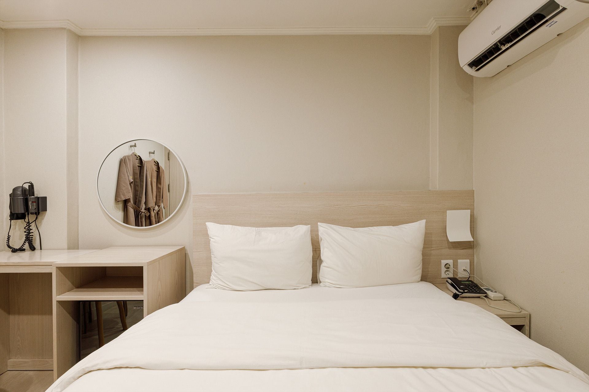 A hotel room with a bed, a desk with a phone, a mirror, and an air conditioning unit.