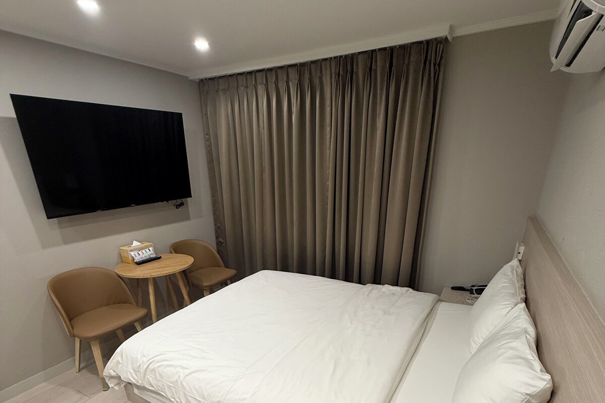 A hotel room with a bed, a TV mounted on the wall, a small table with a chair, and a window with curtains.