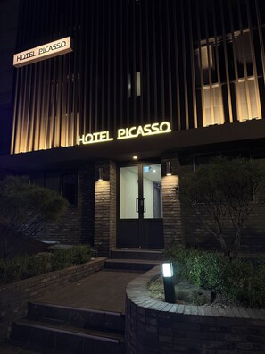 Entrada de hotel com design moderno, destacando uma placa iluminada que diz 'HOTEL PICASSO'.