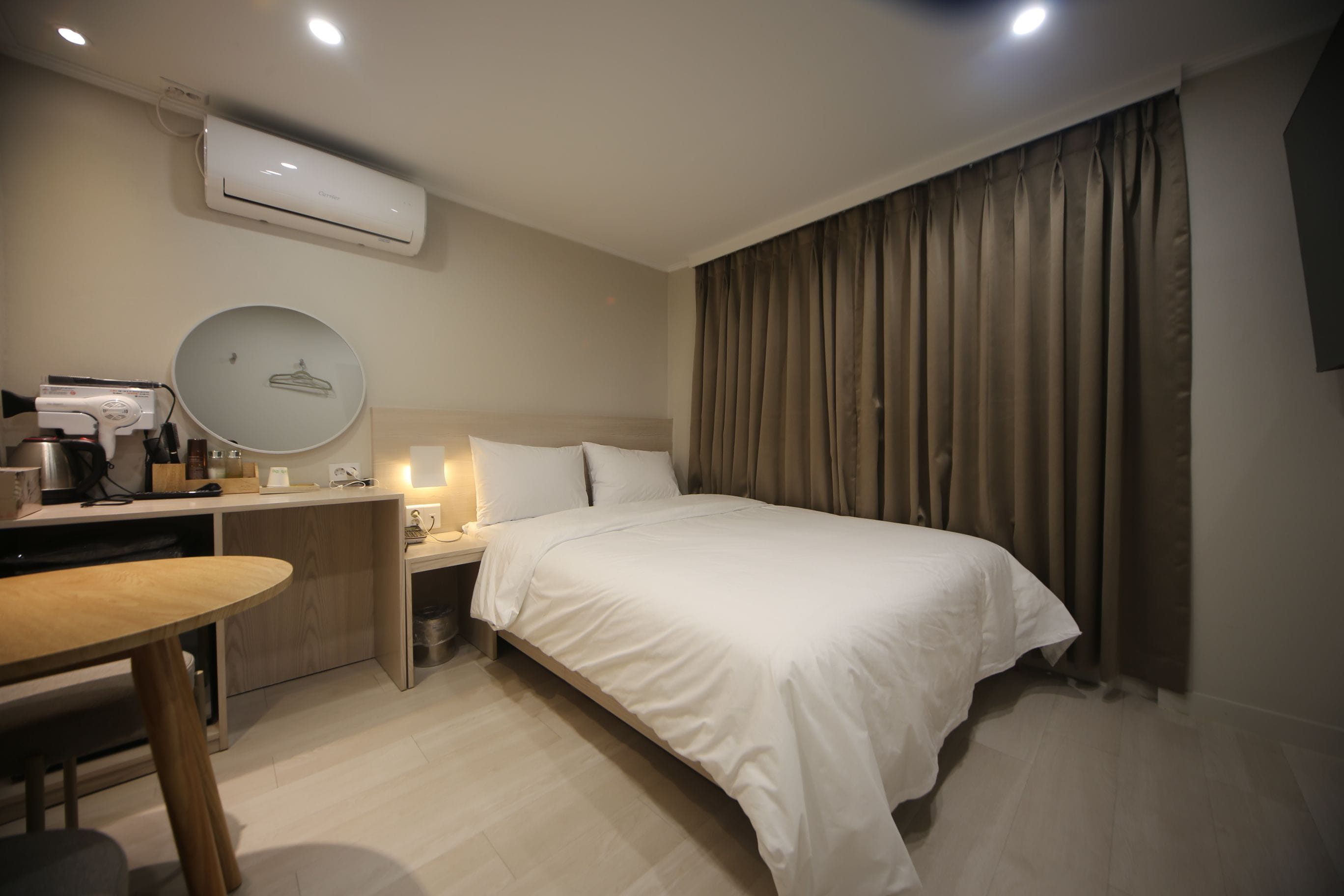 Quarto de hotel com cama, mesa de cabeceira, escrivaninha e aparelho de ar condicionado.