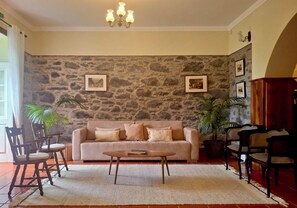 A living room with a stone wall, a beige sofa, a coffee table, and two chairs. - Villa da Fajã Alta - Room No. 4 (Santana)