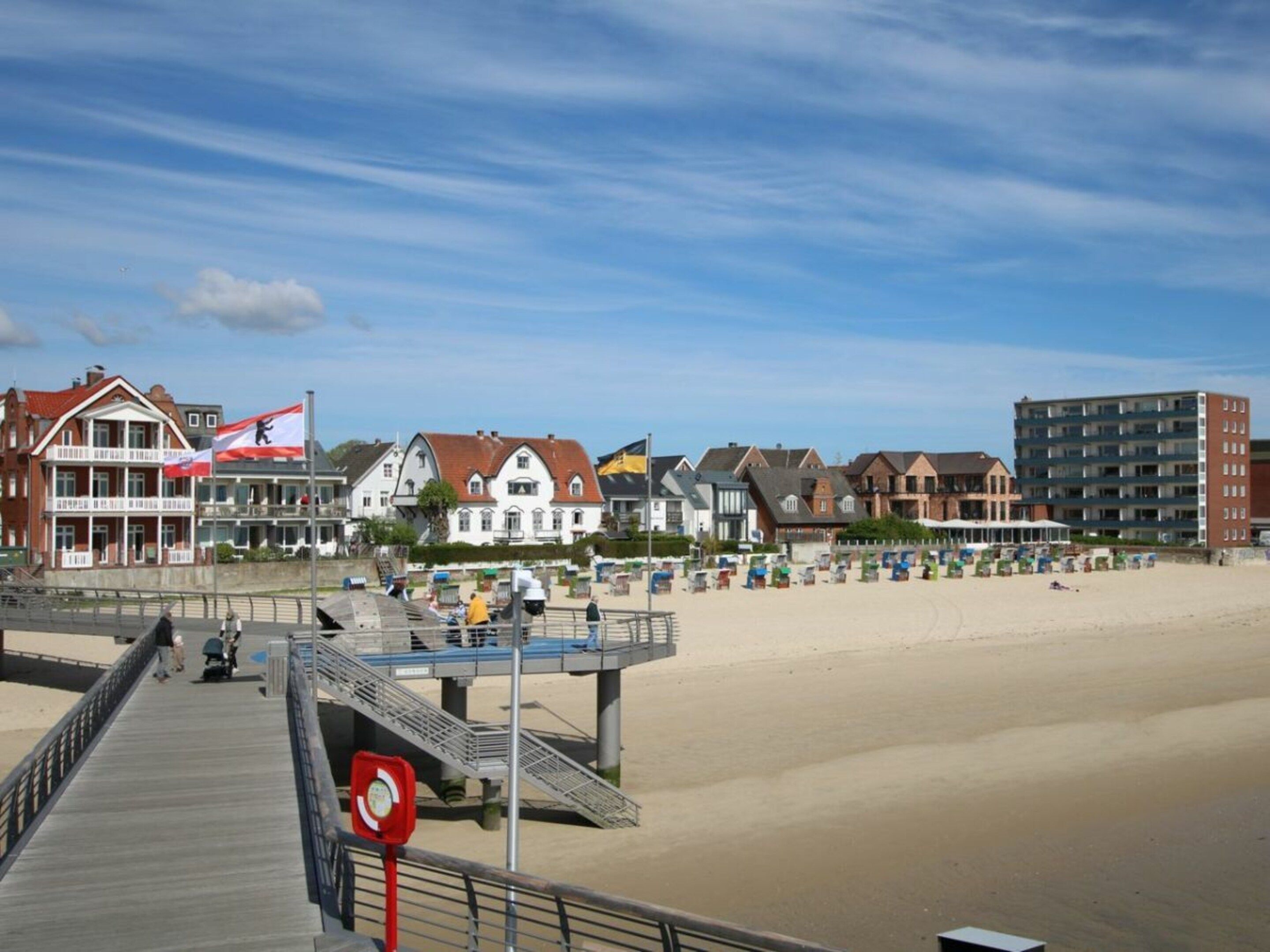 A coastal promenade with a wooden walkway, beach umbrellas, and a variety of buildings including a hotel with a flag.