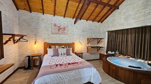 Ein Schlafzimmer mit einem großen Bett, einer Holzbalkendecke, einer Ziegelwand, einem Gemälde, einem Nachttisch, einer Lampe, einem Nachttischregal, einem Holzboden und einer Badewanne.