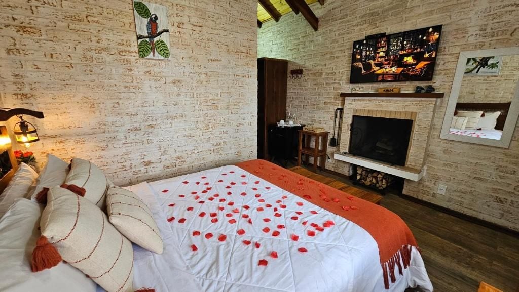 Quarto aconchegante com parede de tijolos, lareira, cama com pétalas de rosa e televisão.