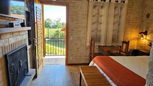 Un dormitorio con cama, cabecera de madera, mesita de noche, ventana con cortinas, chimenea y balcón.