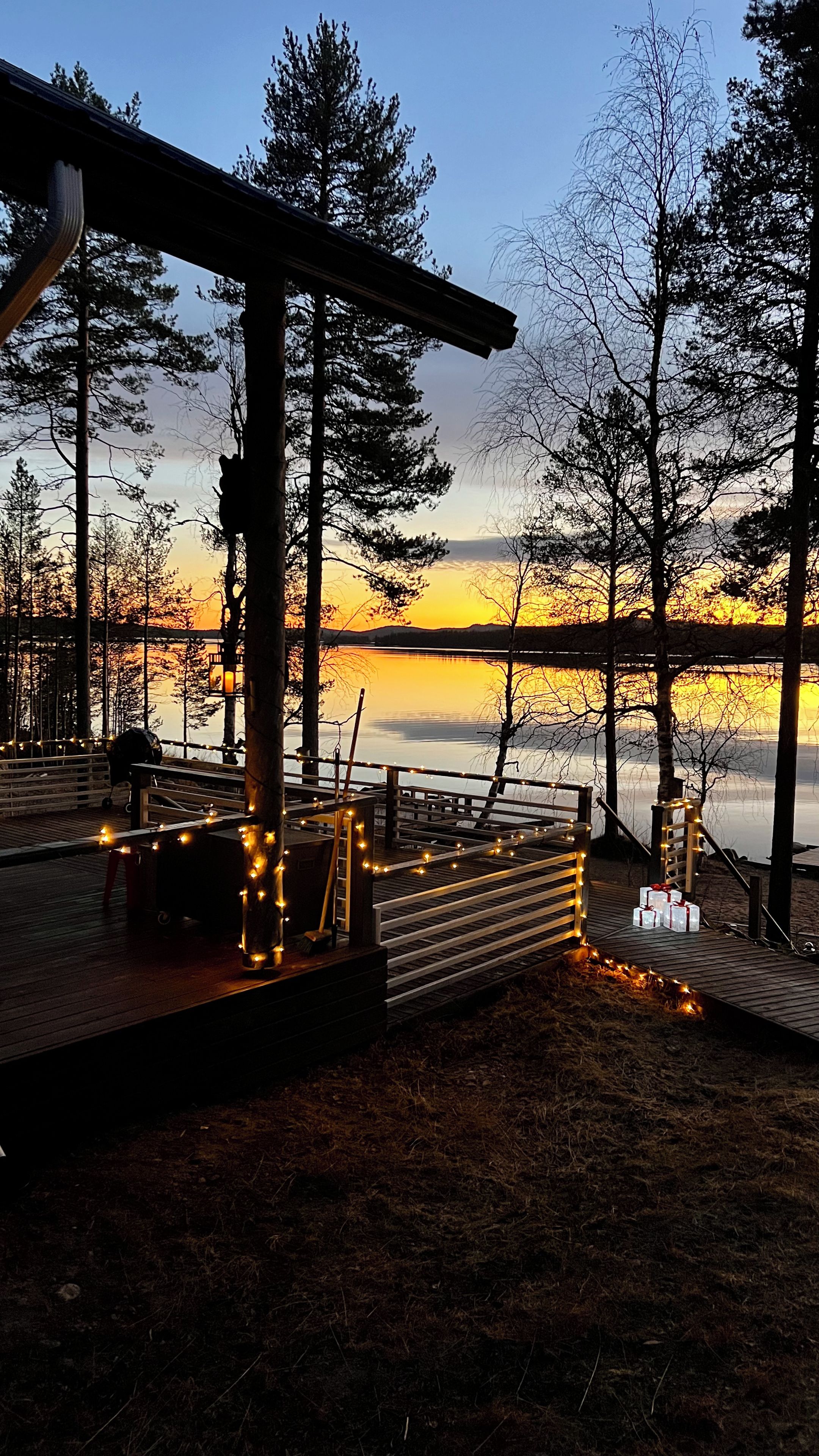 A wooden deck with railings, a bench, and a stroller, overlooking a body of water and trees during sunset.