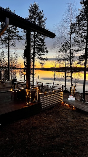 Una terraza de madera con barandillas, un banco y un cochecito de bebé, con vista a un cuerpo de agua y árboles al atardecer.