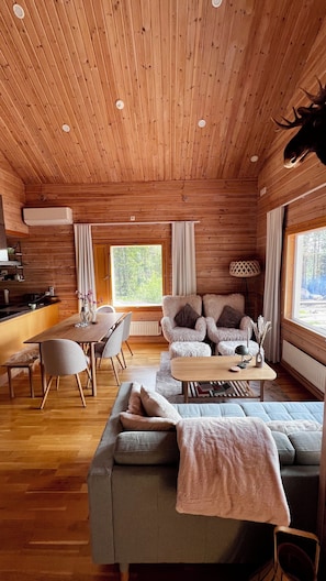 A cozy cabin interior with a wooden ceiling, a dining area, a sofa, and a large window offering a view of the outdoors.