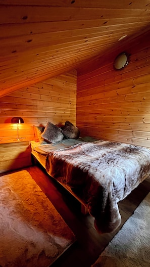Una habitación cálida tipo cabaña con techo de madera, una cama con manta, almohadas y una lámpara de noche.