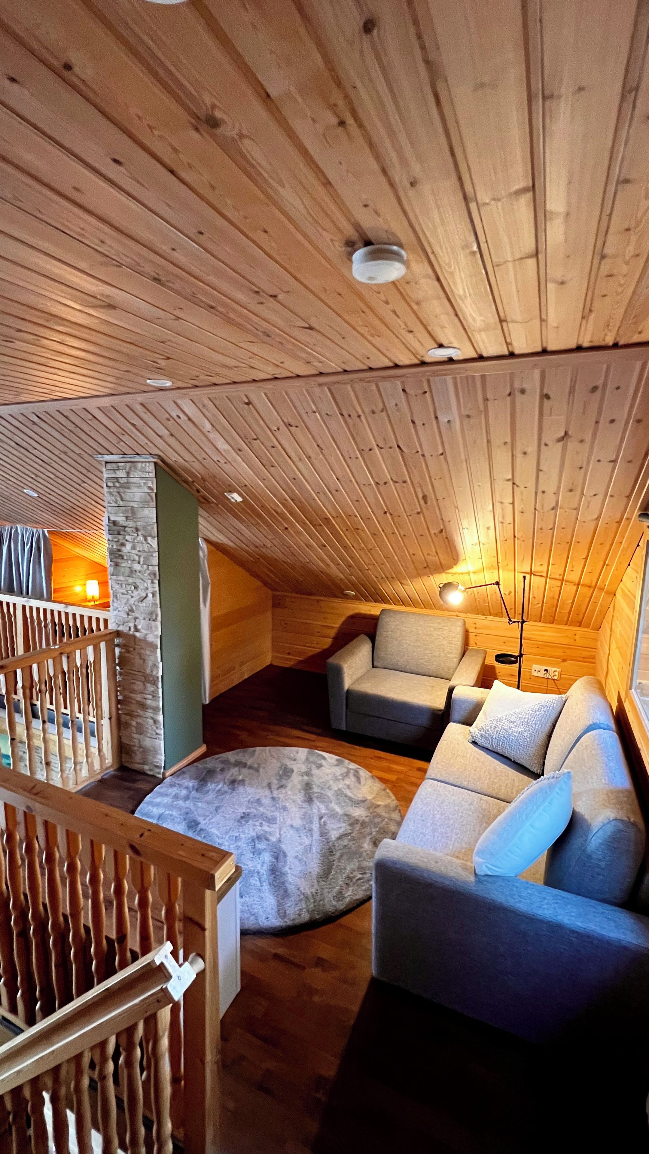 A cozy attic room with wooden beams, a stone wall, a cushioned sofa, and a wooden railing.