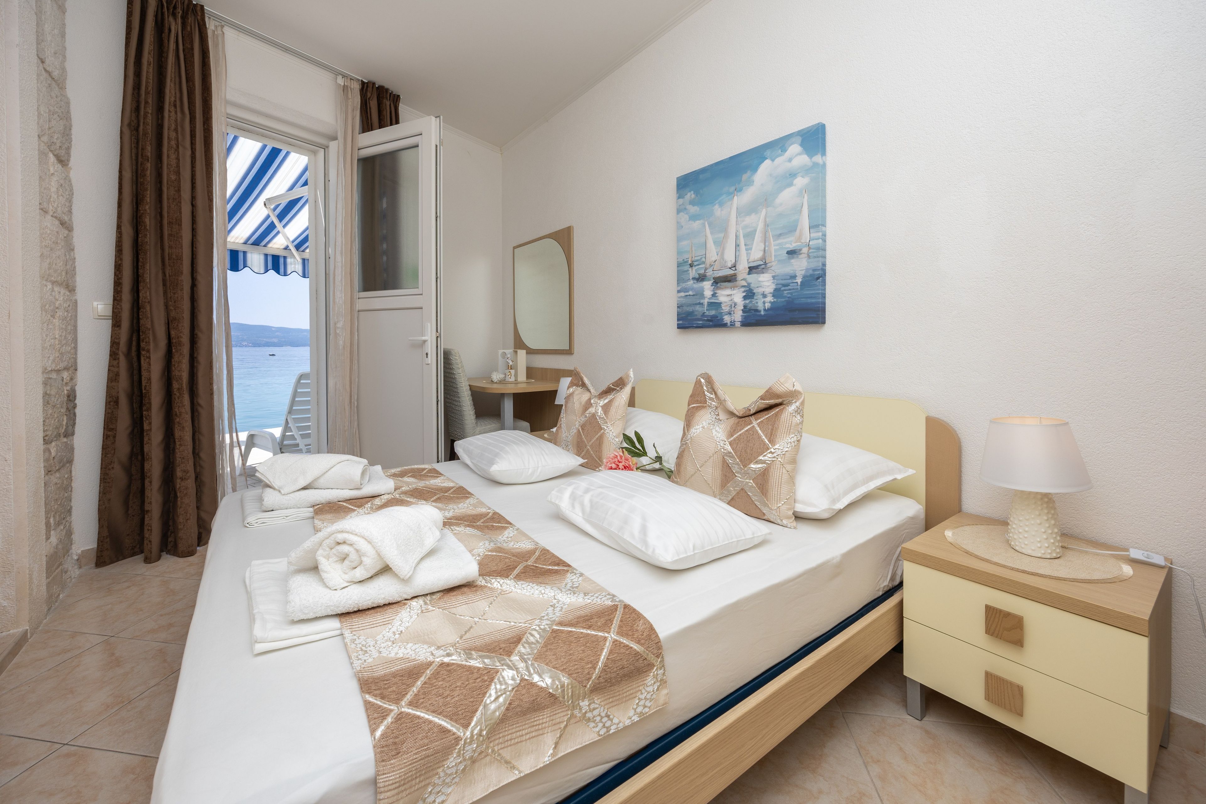 A hotel room with a bed, a bedside table with a lamp, a painting on the wall, and a view of the sea through the window.