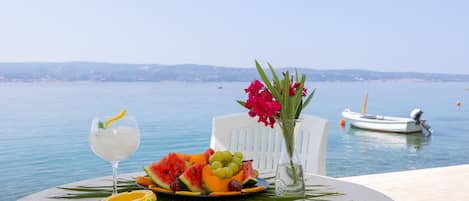 Una mesa con una bebida, fruta y flores junto a un cuerpo de agua con barcos al fondo.
