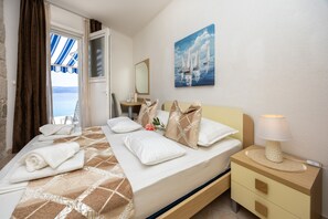 Habitación de hotel con una cama, una mesita de noche con lámpara, un cuadro en la pared y vista al mar a través de una ventana.