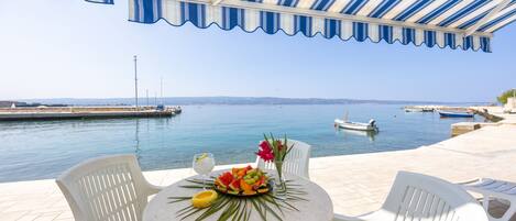 A terrace with a view of the sea, featuring a table with fruit and flowers, white chairs, and a striped awning.