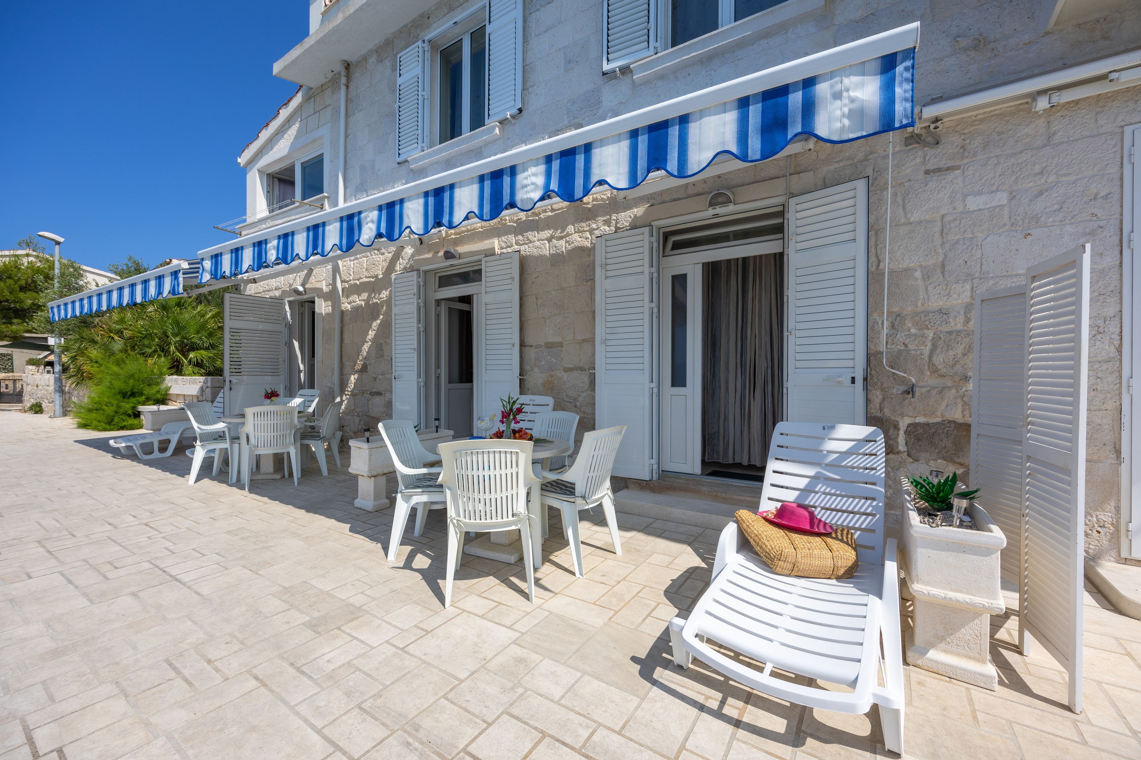 Un bâtiment à la façade en pierre, avec des volets blancs et un auvent bleu. On y trouve des espaces de détente en plein air, avec des chaises et des tables en plastique blanc.
