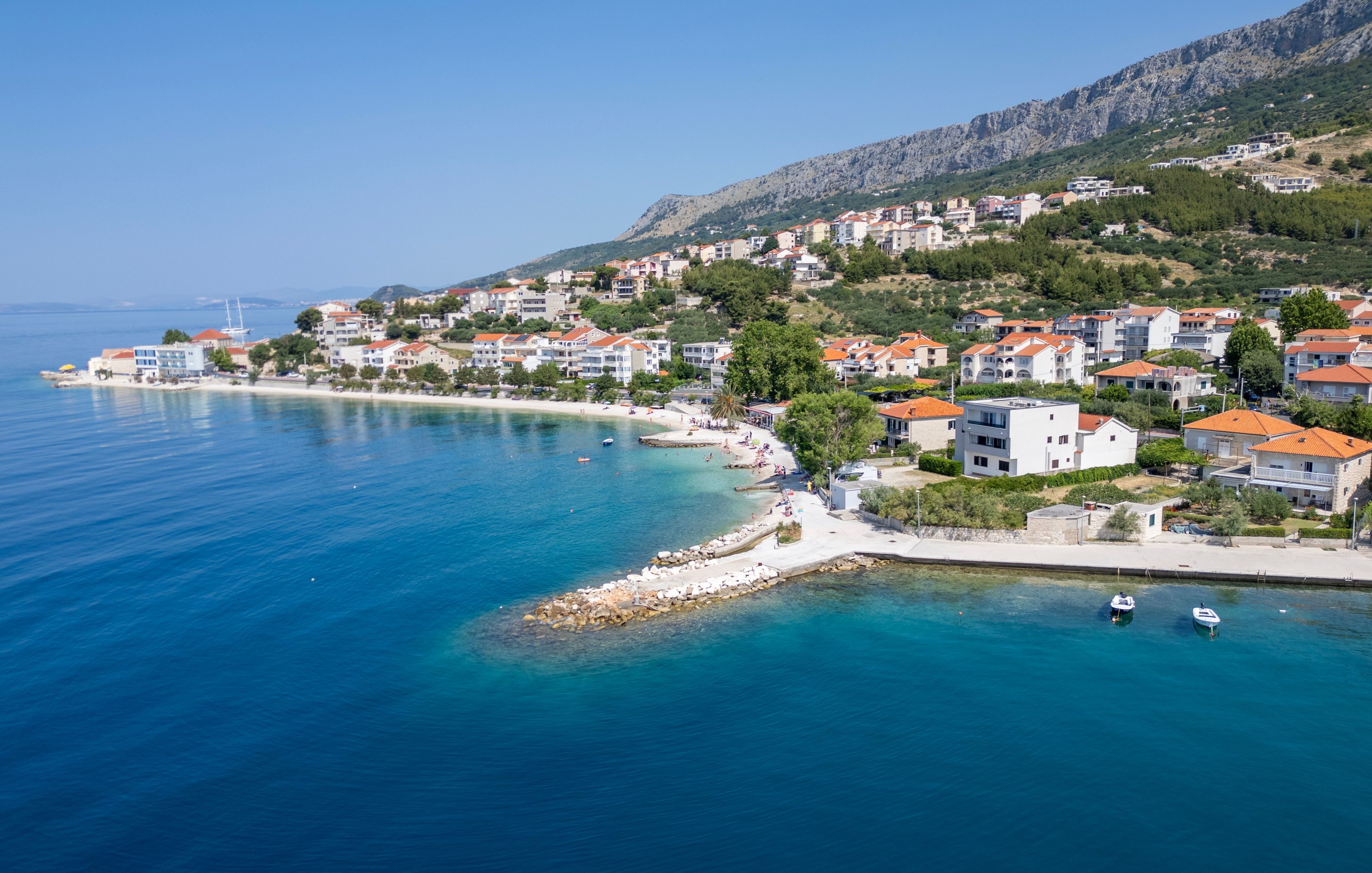 Une ville côtière avec des bâtiments qui bordent le front de mer, une mer d’un bleu limpide et un paysage montagneux en arrière-plan.