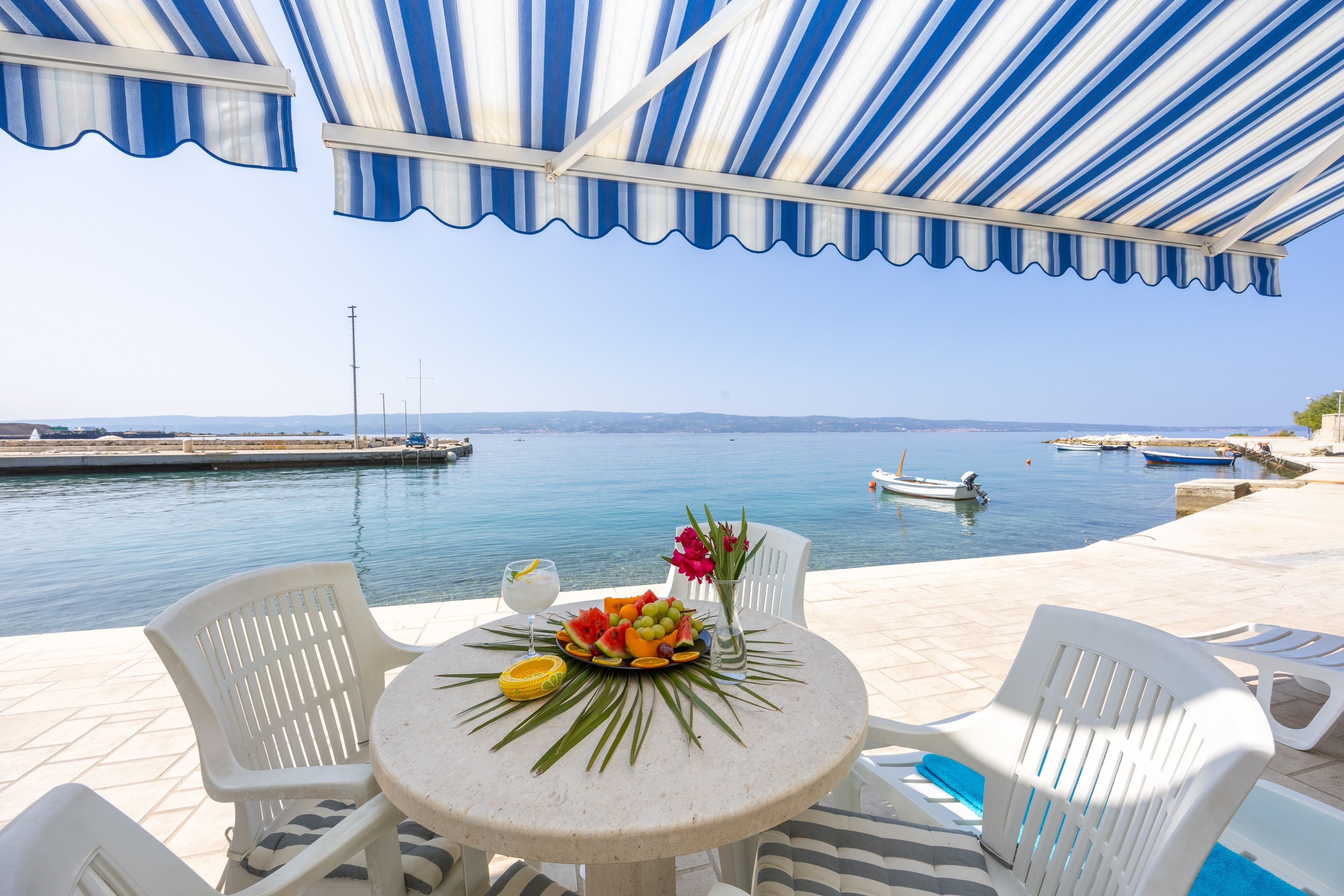 A terrace with a view of the sea, featuring a table with fruit and flowers, white chairs, and a striped awning.
