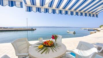 A terrace with a view of the sea, featuring a table with fruit and flowers, white chairs, and a striped awning.