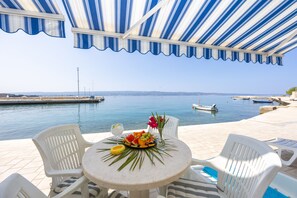 A terrace with a view of the sea, featuring a table with fruit and flowers, white chairs, and a striped awning.
