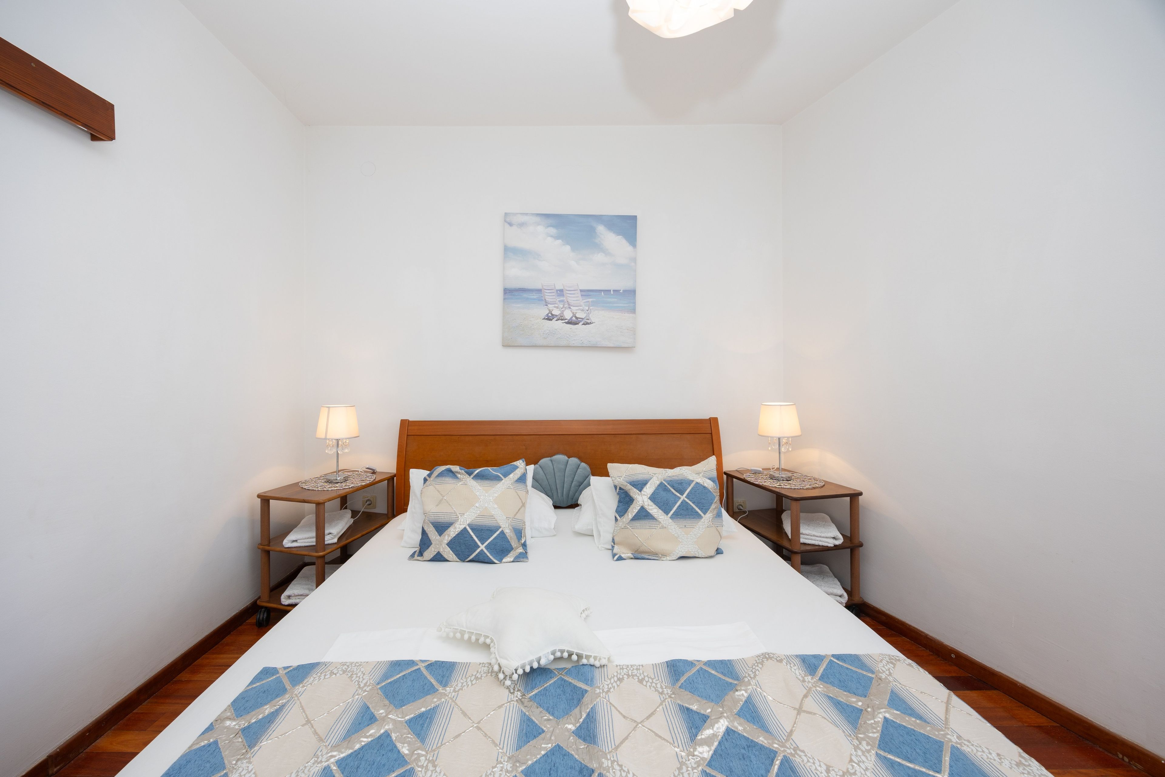 Un dormitorio con cabecera de madera, dos mesitas de noche con lámparas, un cuadro de una escena de playa y una colcha estampada.
