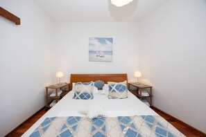 Un dormitorio con cabecera de madera, dos mesitas de noche con lámparas, un cuadro de una escena de playa y una colcha estampada.
