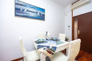 A dining area with a table set for two, white chairs, a painting of sailboats on the wall, and a door with an emergency exit sign.