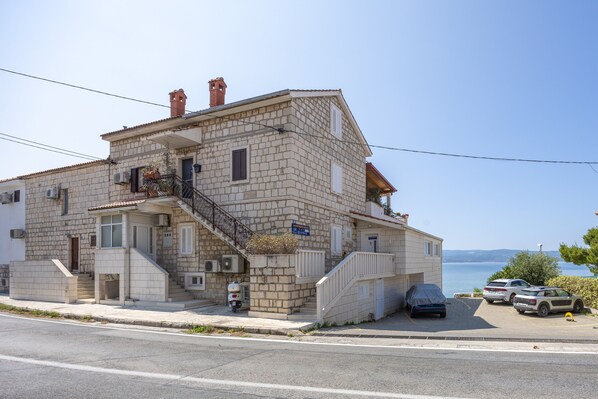 A two-story building with a stone facade, a staircase leading to the entrance, and a parking area with cars.