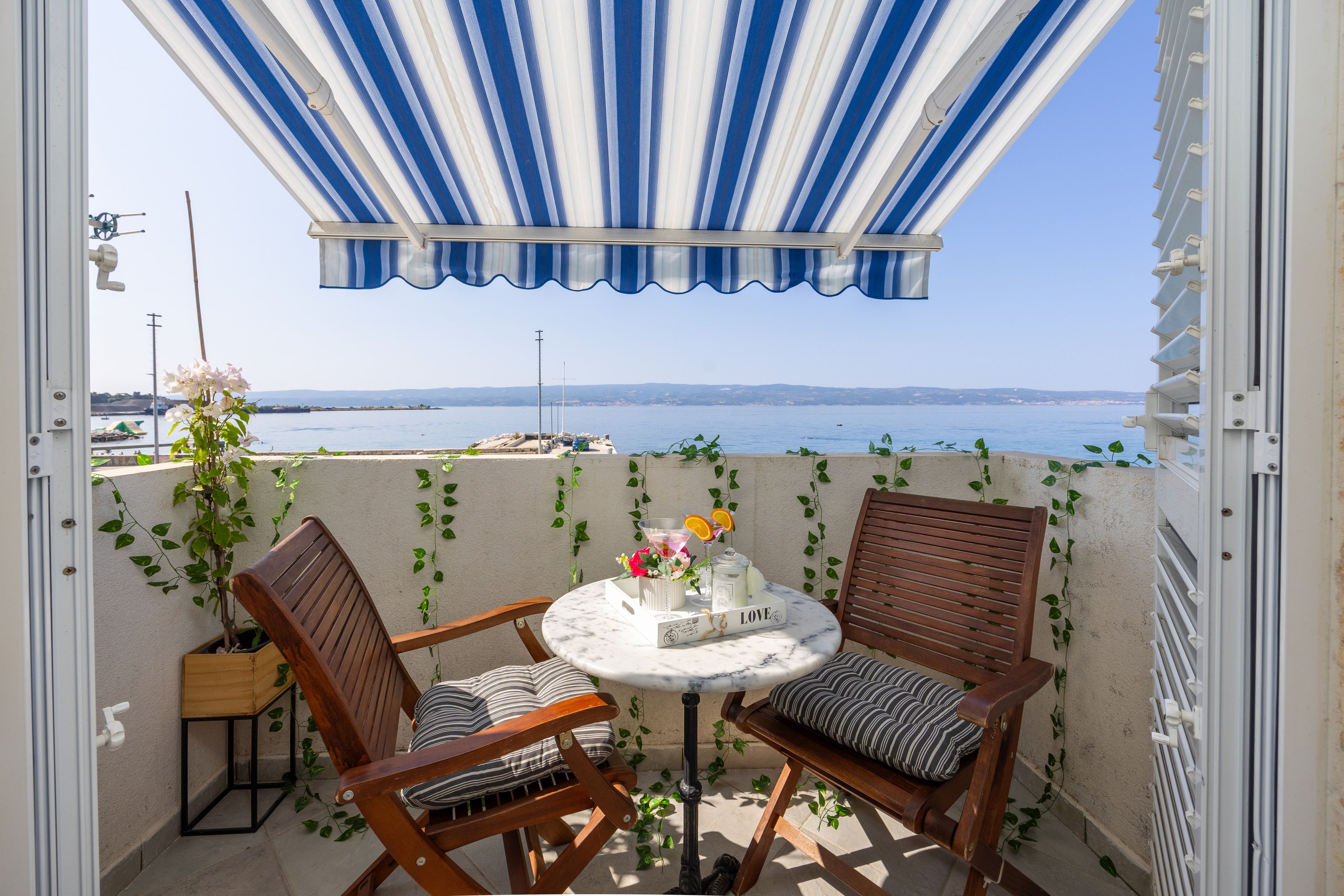 Un balcon avec vue sur la mer, comprenant deux chaises en bois, une petite table ronde et un auvent à rayures.