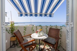 A balcony with a view of the sea, featuring two wooden chairs, a small round table, and a striped awning.
