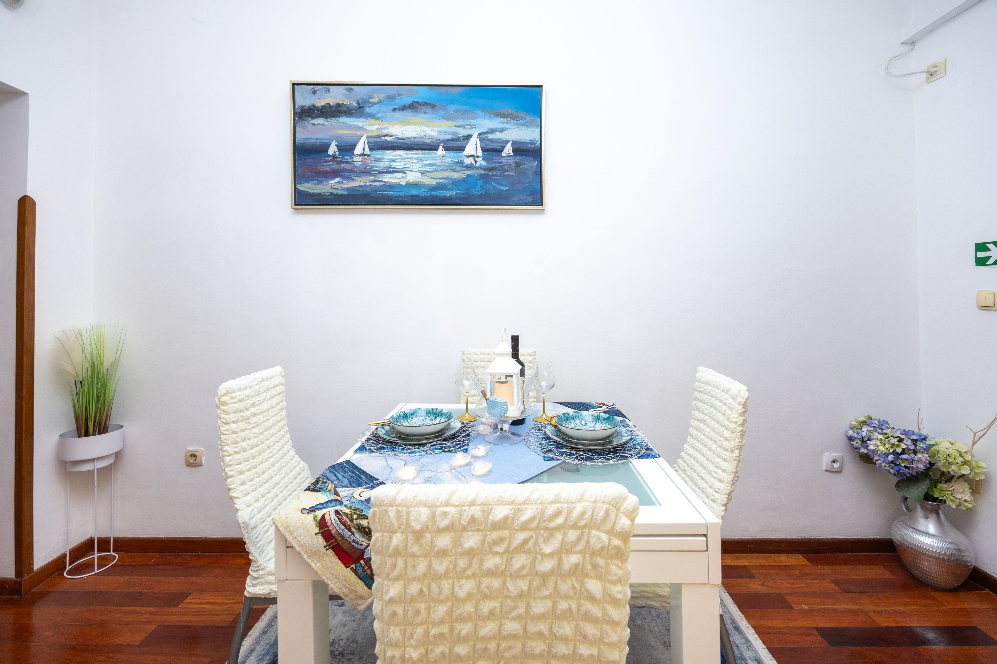Een eetruimte met een tafel gedekt voor vier personen, een schilderij erboven en een potplant aan de linkerkant.