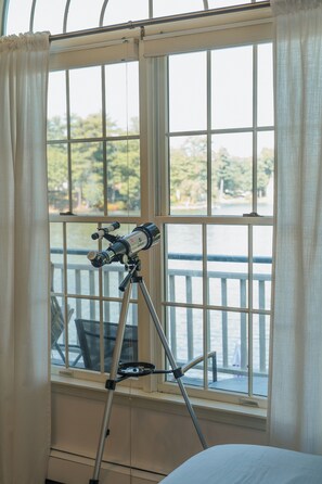 A telescope on a tripod by a window with a view of a body of water and trees.