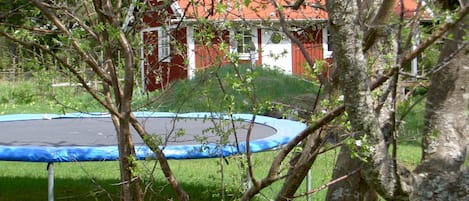 A trampoline in a grassy area with trees and a red-roofed building in the background.