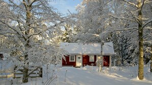 Uma cabana coberta de neve, com fachada vermelha, cercada por árvores e um muro de madeira.