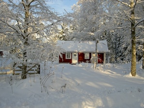 A snow-covered cabin with a red exterior, surrounded by trees and a wooden fence.