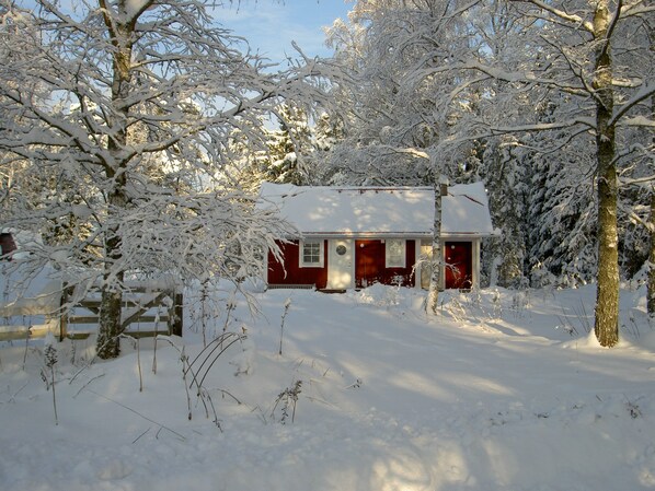 Una cabaña nevada con fachada roja, rodeada de árboles y una cerca de madera.