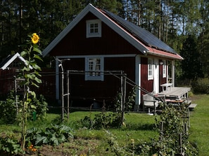 A red wooden house with a sloped roof, a small porch, and a fenced garden area.