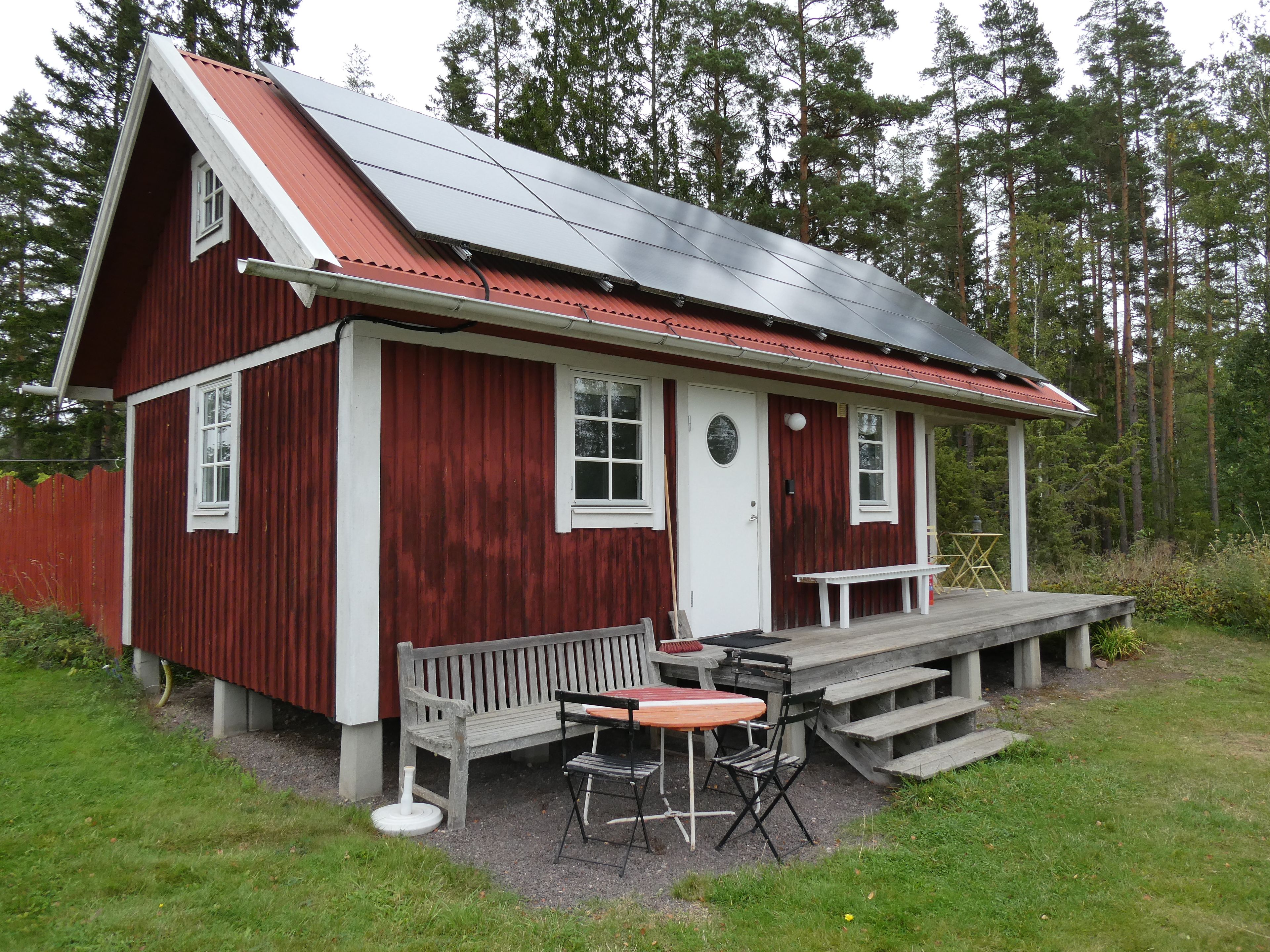 A red wooden building with a sloped roof, a front porch, and a small outdoor seating area with a table and chairs.