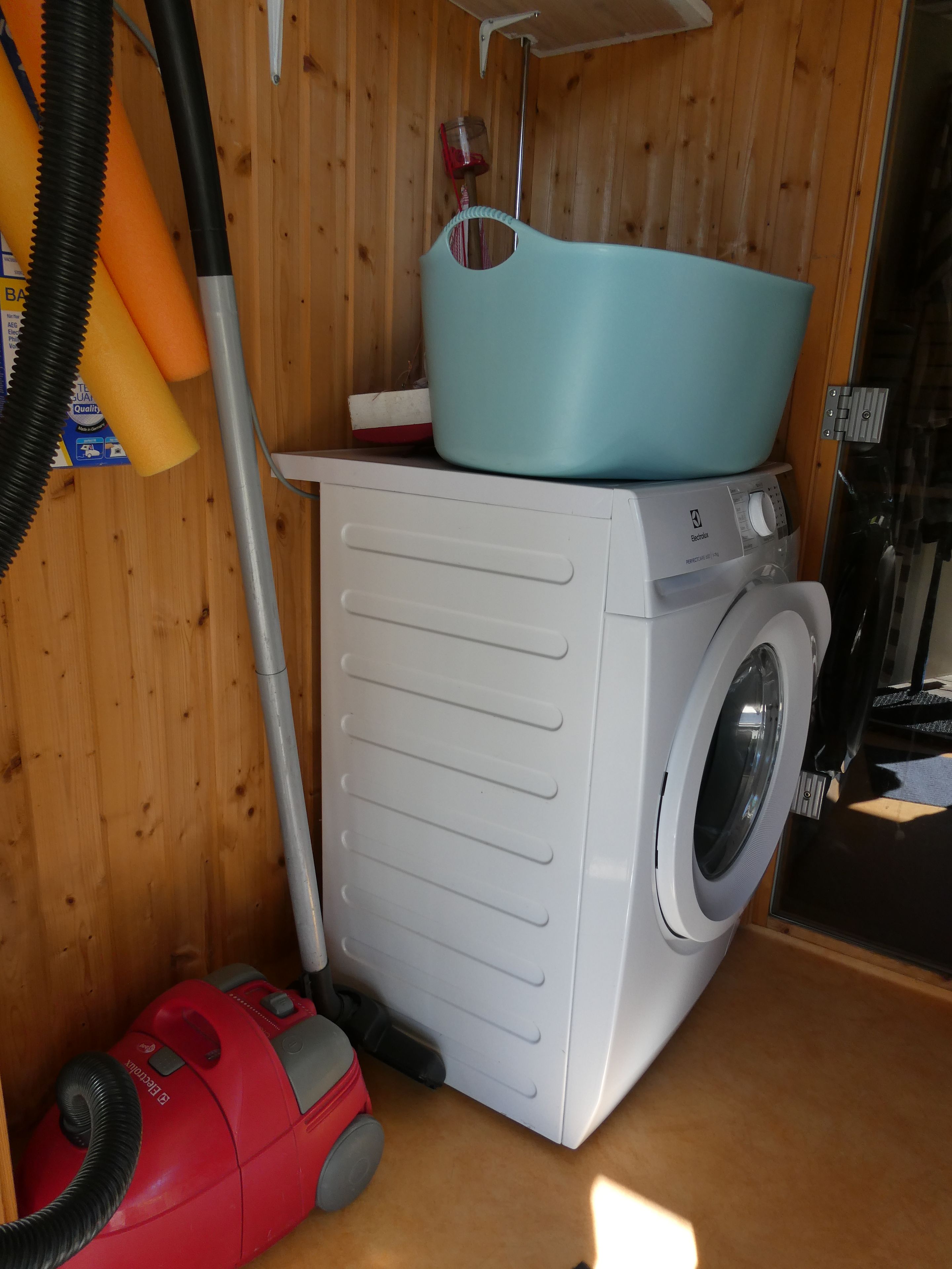 Un espace réduit dédié à la buanderie, comprenant un lave-linge, un aspirateur et un évier bleu posé sur un meuble en bois.