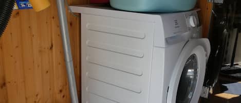 Een kleine wasruimte met een wasmachine, een stofzuiger en een blauwe wasbak op een houten kastje.