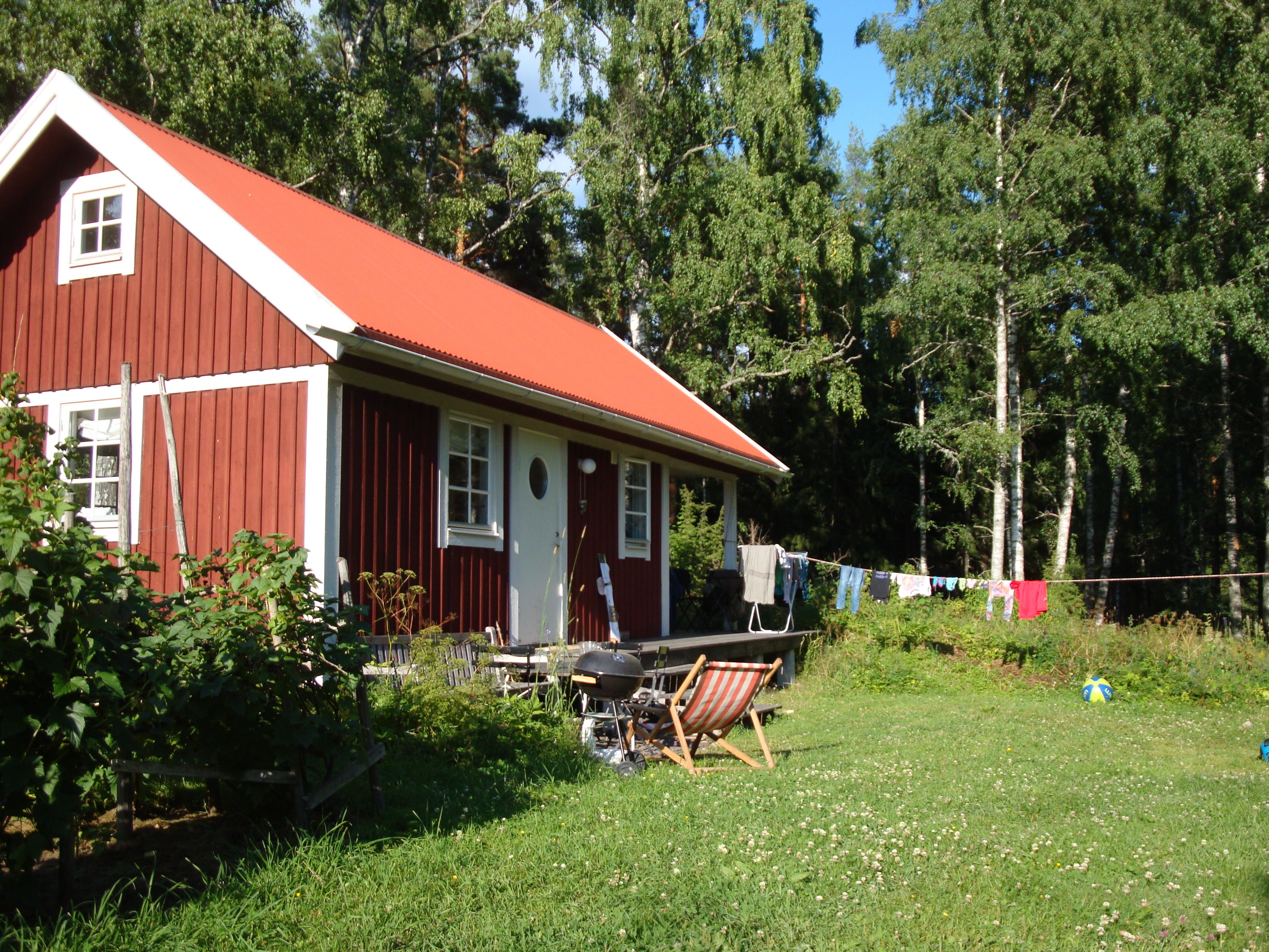 Ett rött trähus med en vit dörr och vita fönster, en grill och en torklina med tvätt som hänger på den.