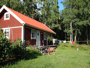 A red wooden house with a white door and windows, a barbecue grill, and a clothesline with laundry.